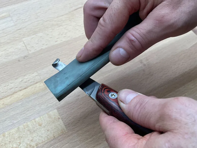 Farrier sharpening a hoof knife with a sharpening stone on a wooden table.