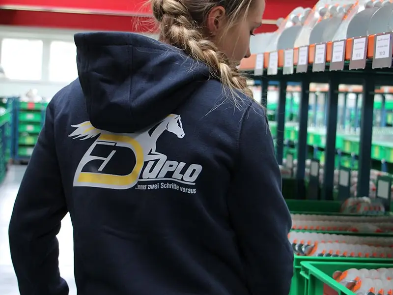 Un’impiegata sta raccogliendo i ferri convenzionali per l’ordine di un cliente nel magazzino; indossa una felpa con cappuccio blu navy di Carhartt.