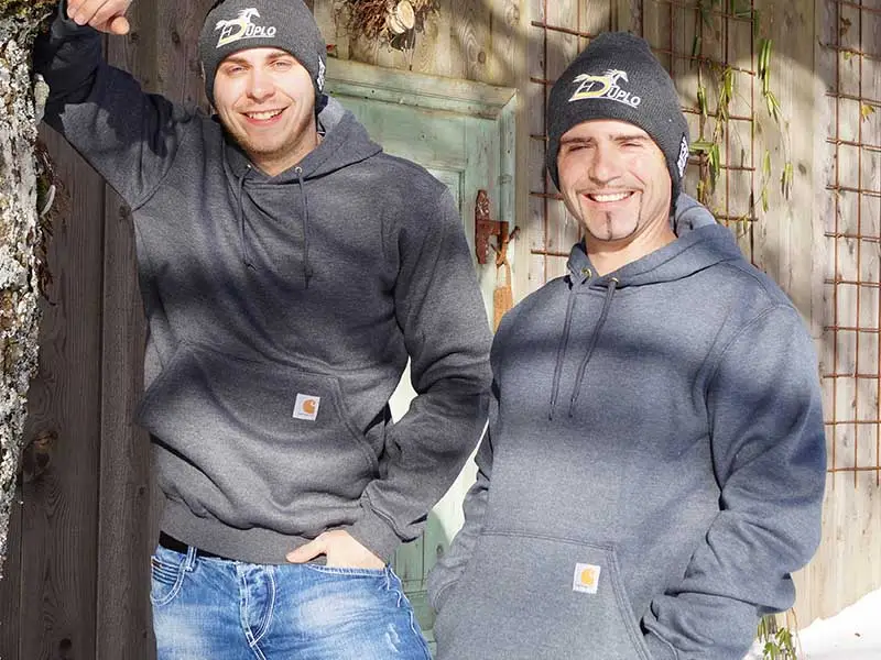 Due uomini indossano la felpa grigia con cappuccio Carhartt davanti a un vecchio edificio di scuderia, il sole splende ma il terreno è ancora coperto di neve. Entrambi indossano anche un berretto di lana.