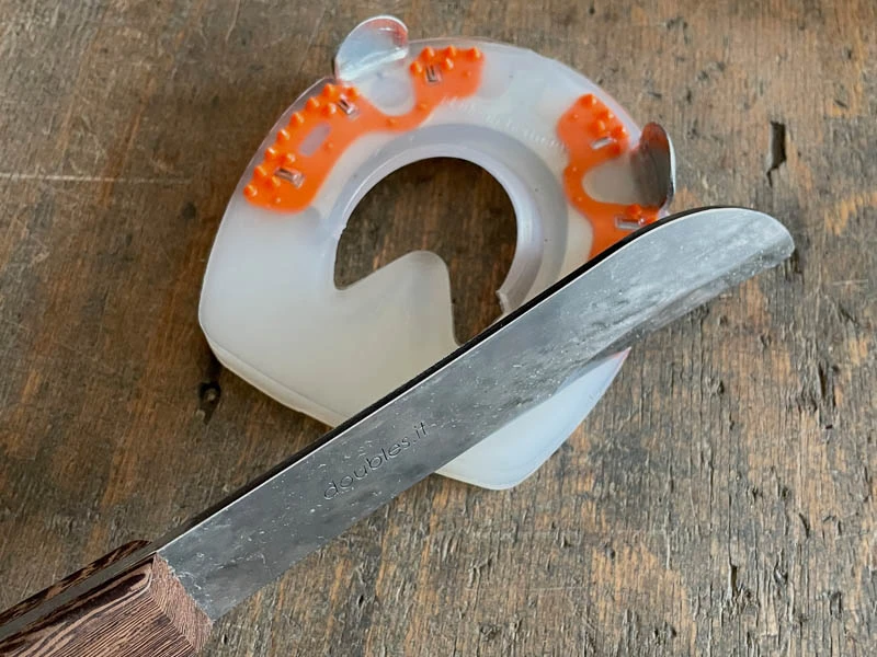 Gros plan sur la lame du rogne-pied Kukri Double S. On voit que la lame doit servir à ajuster le revêtement en plastique d’un fer composite pour chevaux. Le fer est posé sur un établi.