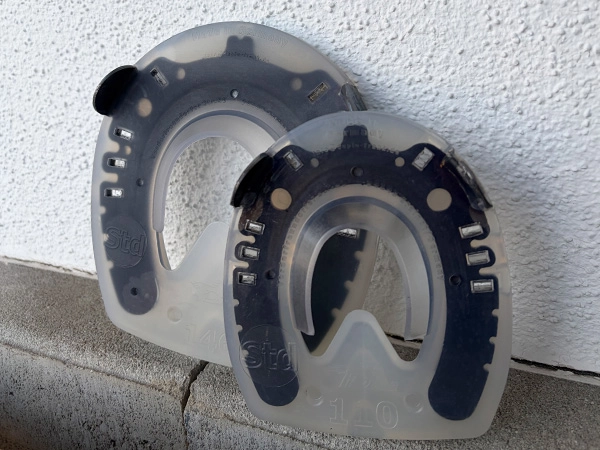 Un Full Branch Shoe avec branche de fer à cheval régulière, offrant davantage de stabilité dans la zone des talons, en taille 110mm, est placé devant un modèle de taille 146mm contre un mur d'écurie.