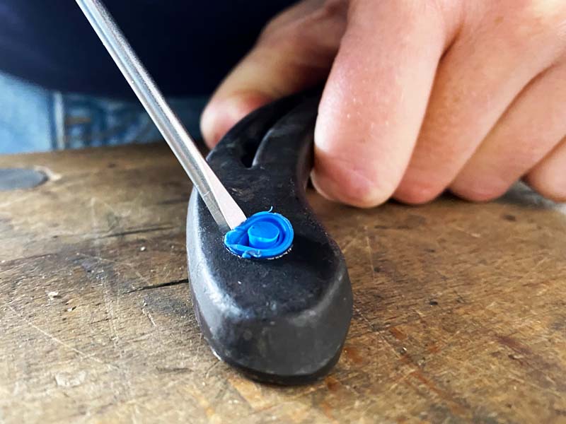 Una mano sostiene una herradura, un destornillador de ranura presiona en el agujero para ramplones para retirar el tapón de cierre azul