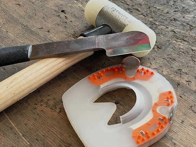 ein Schonhammer mit Nylonkopf, eine Hauklinge in Beilform mit schwarzem ergonomischen Griff und ein stoßdämpfendes Hufeisen liegen auf einer Werkbank