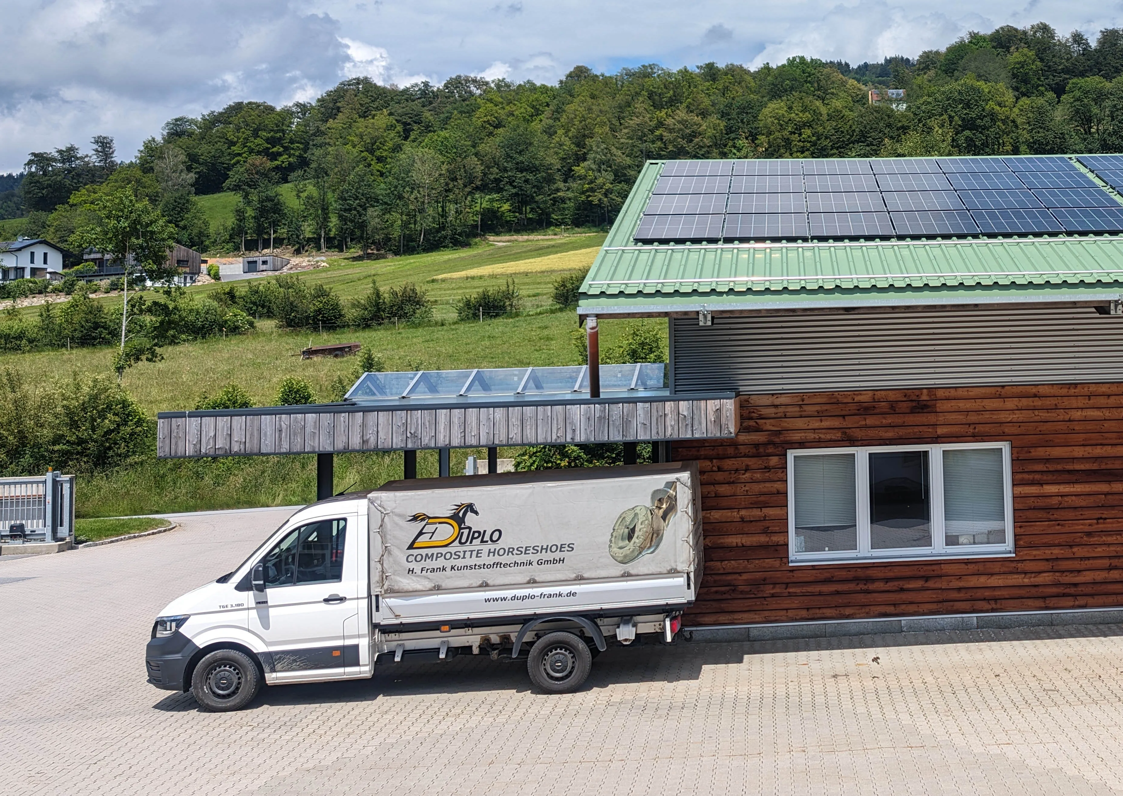 View of the company building of the manufacturer of composite horseshoes in the Bavarian Forest. A company vehicle with logo and part of the production facility are visible. A photovoltaic system is installed on the roof.