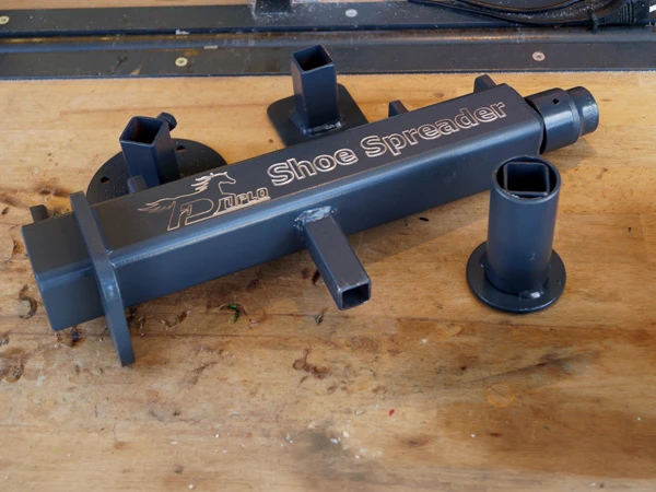 Der Duplo Hufeisenspreizer, der sogenannte Shoe Spreader, liegt auf einer Werkbank. Daneben liegen drei Adapter, mit denen der Spreizer auf der Werkbank, am Amboss oder am Hufbock genutzt werden kann.