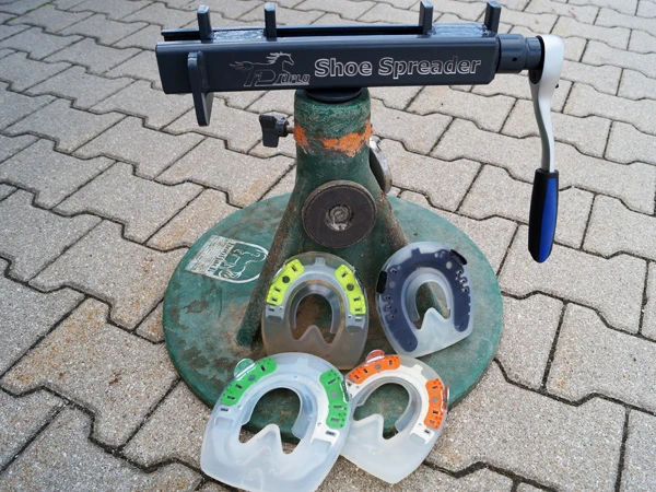 Der Duplo Shoe Spreader ist in einem grünen Hoofjack® von Kerckhaert montiert. Davor liegt eine Auswahl an Verbundhufeisen, die mit dem Spreizer enger oder weiter geformt werden können.