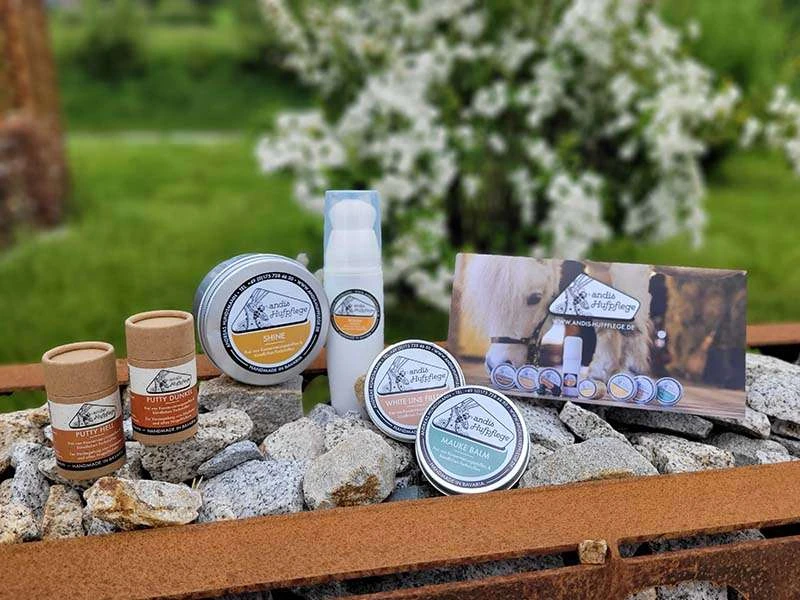 Putty, Shine, Polish, Mauke Balm und White Line Filler von andis Hufpflege stehen auf Steinen neben dem Produktflyer