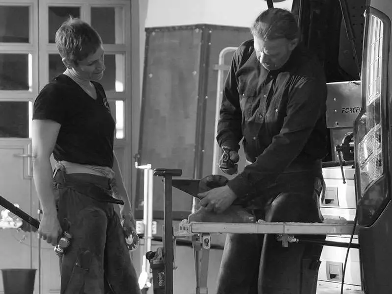 A farrier instructor explains to a farrier how to fit an alternative horseshoe with a hammer. He beats the horseshoe into the right shape on the anvil.