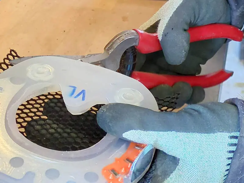 Un maniscalco tiene in mano un ferro convenzionale rivestito in materiale plastico e una cesoia per cuoio con impugnatura rossa. Indossa guanti antitaglio. Sotto il ferro si vede la rete rigida chiamata MESH di glue-u, che sta per essere tagliata.