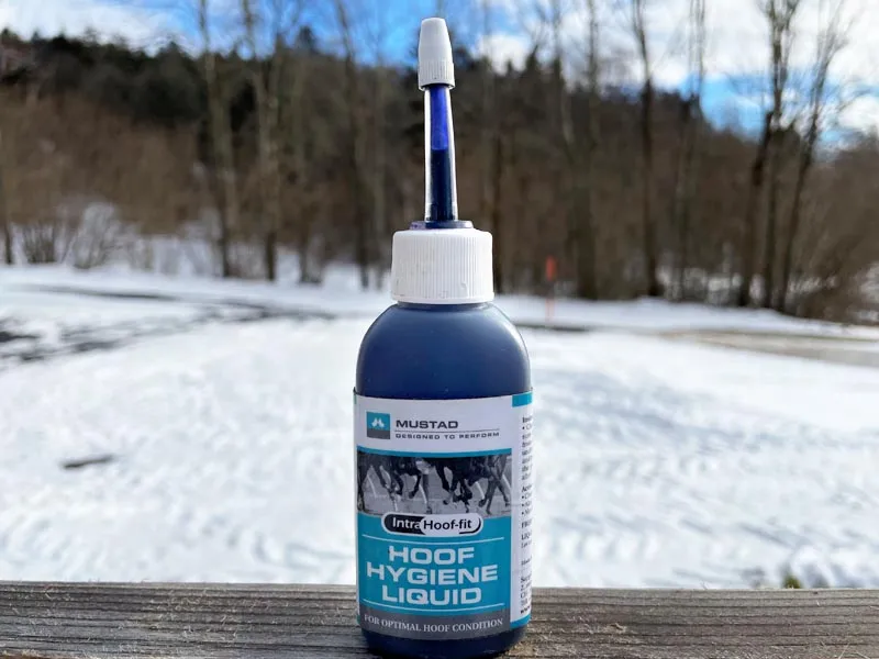 Das Hoof Hygiene Liquid von Mustad steht auf einem Holzbalken. Im Hintergrund ist eine winterliche Landschaft zu sehen.