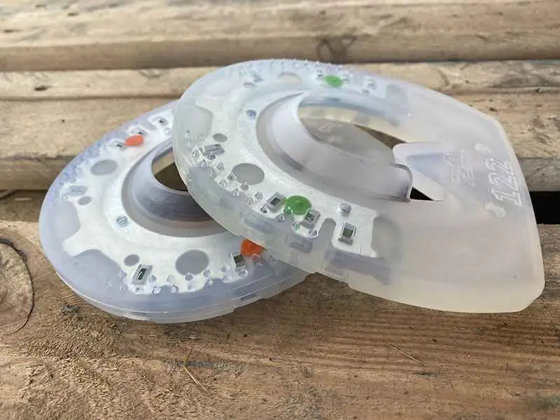 Zwei Kunststoff-Hufeisen mit Metallkern liegen auf einer Holzoberfläche. Eines ist mit grünen Punkten markiert und kennzeichnet die härtere Extra-Variante, das andere mit orangefarbenen Punkten steht für die Standardversion aus etwas weicherem Kunststoff.