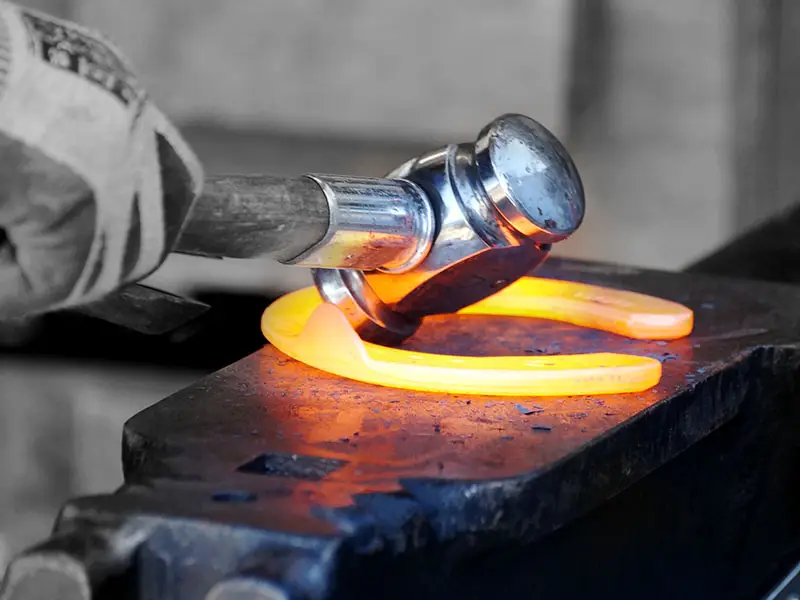 The farrier is shaping a hot horseshoe on the anvil. He wears protective gloves and uses a forging hammer by Double S.