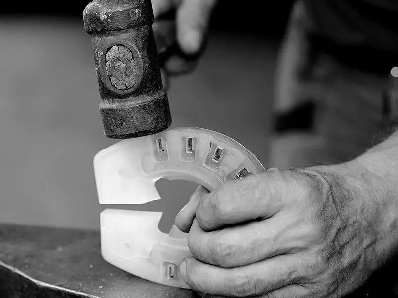 A modified composite horseshoe is held upright on the anvil by a farrier. In the other hand, a forging hammer is used to make adjustments.