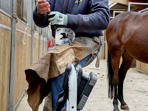 Un herrador modifica una herradura compuesta. Utiliza el delantal de herrar como apoyo sobre su muslo.