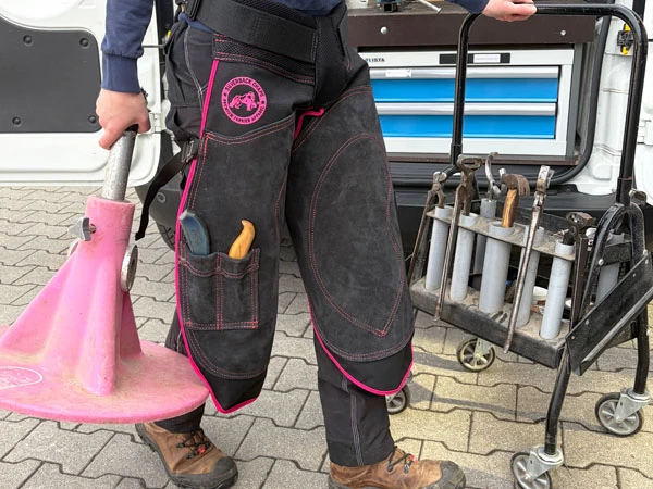 Una herradora lleva el delantal Silverback Chapette con costuras rosas. En la mano derecha sostiene un Hoofjack® rosa y con la izquierda mueve su carro de herramientas hacia la zona de herraje.